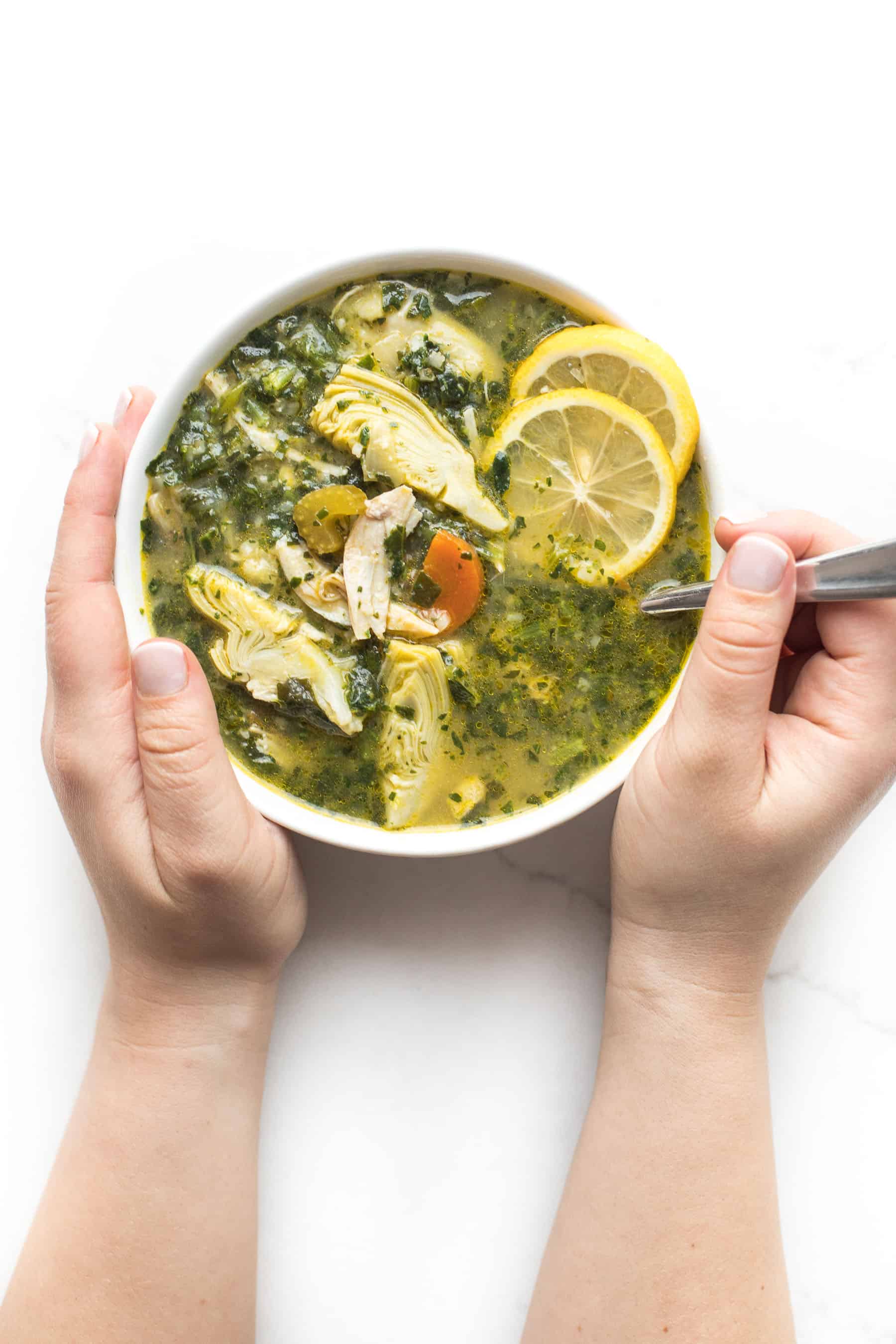 2 hands holding lemon artichoke chicken soup in a white bowl