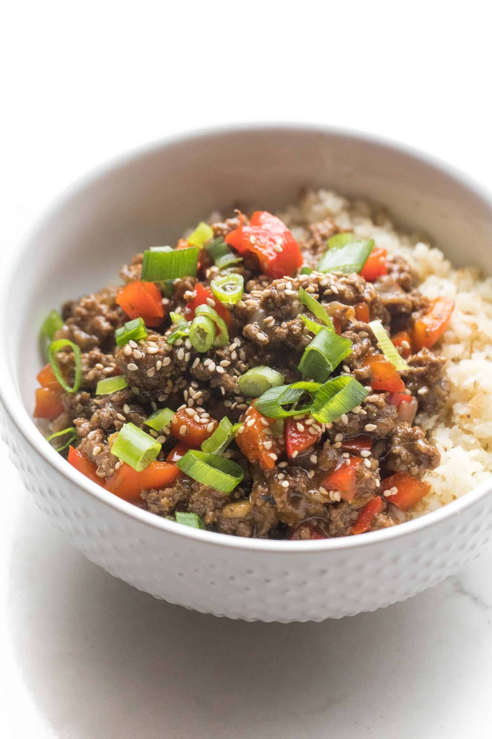 ground asian beef + bell peppers topped with green onions over cauliflower rice in a white bowl