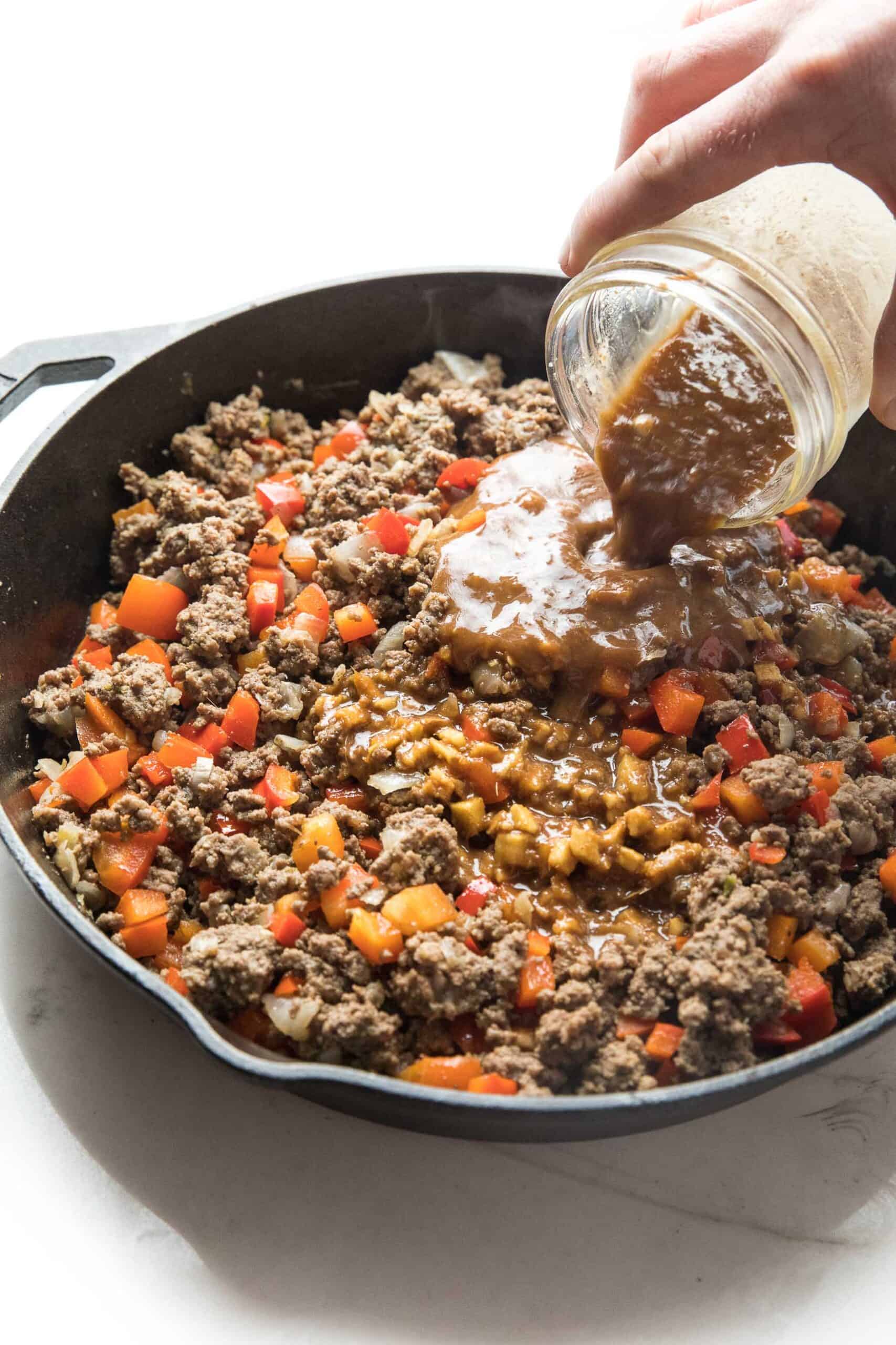 pouring sauce over ground beef and bell peppers in a cast iron skillet