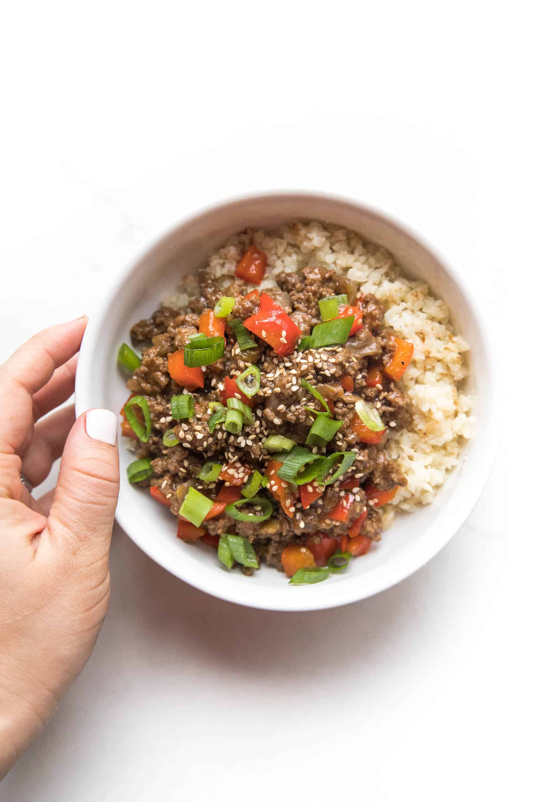 hand holding ground asian beef + bell peppers topped with green onions over cauliflower rice in a white bowl