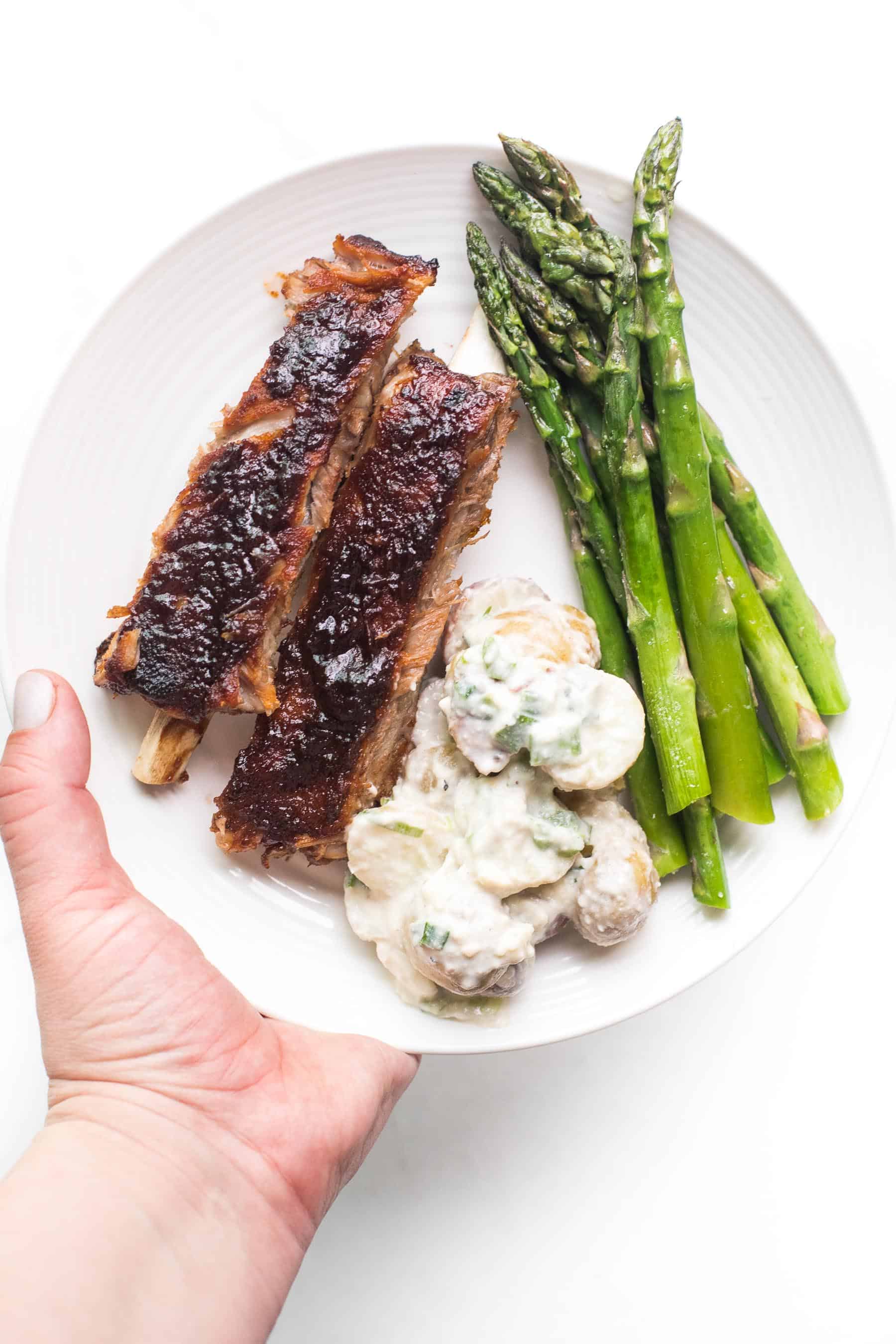 hand holding a white plate with ribs, asparagus and potato salad on a white background