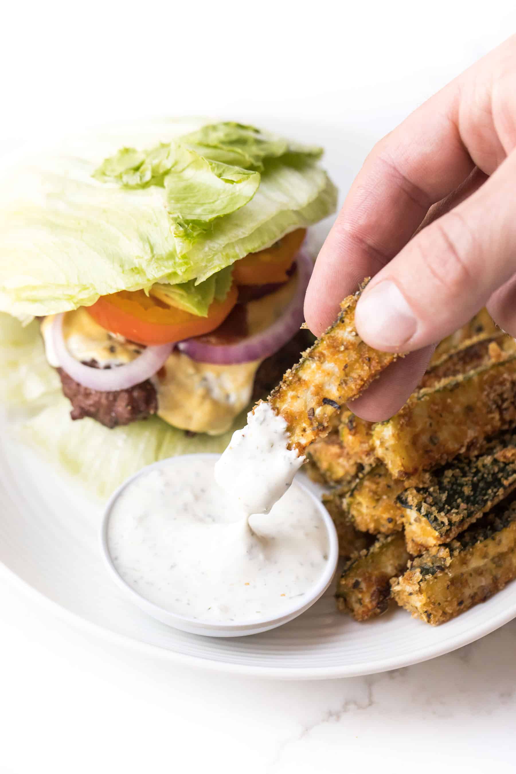 Hand dipping keto zucchini fries in ranch dressing on a white plate and white background with a lettuce wrapped burger on the plate