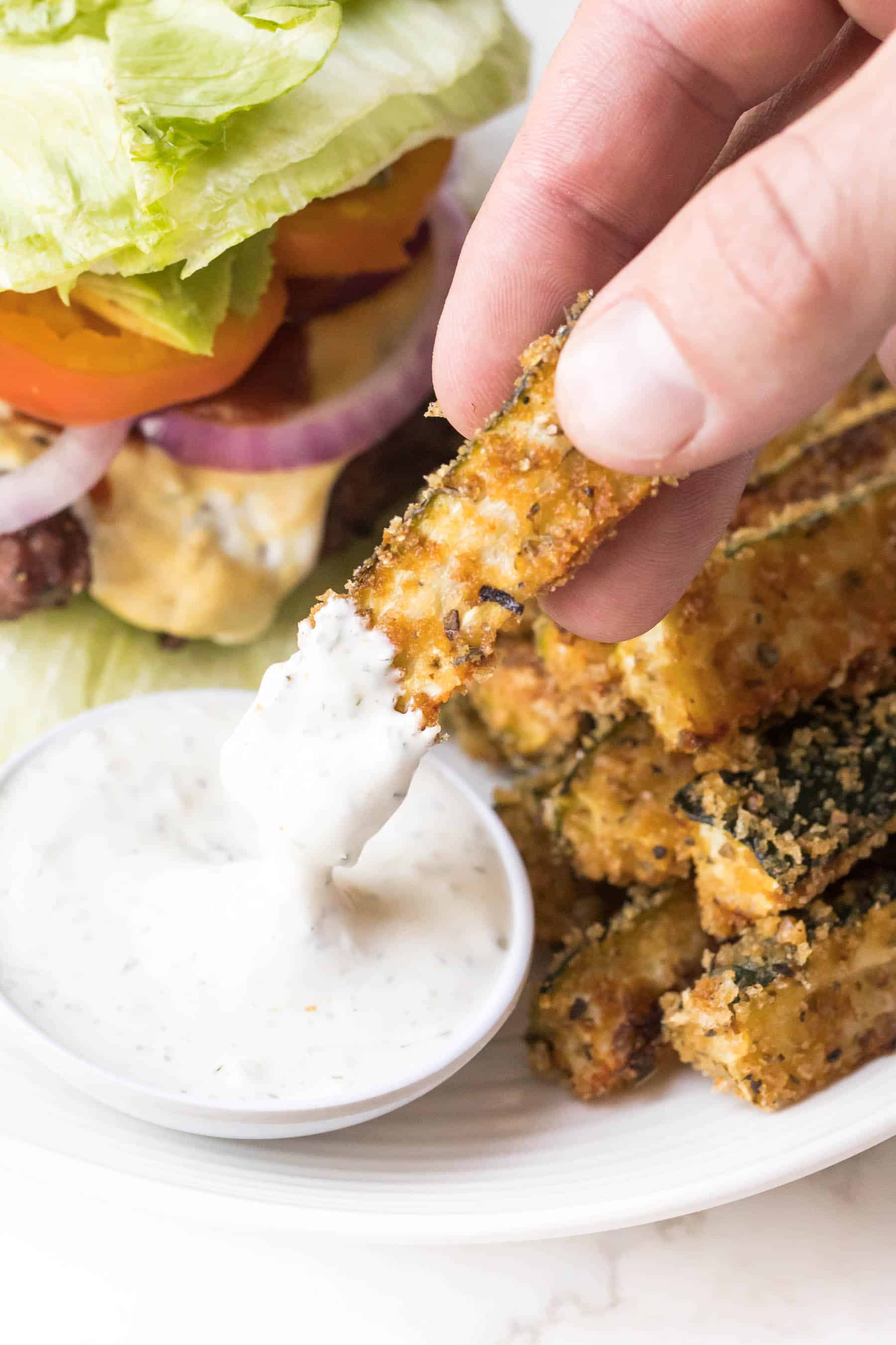 Hand dipping keto zucchini fries in ranch dressing on a white plate and white background with a lettuce wrapped burger on the plate