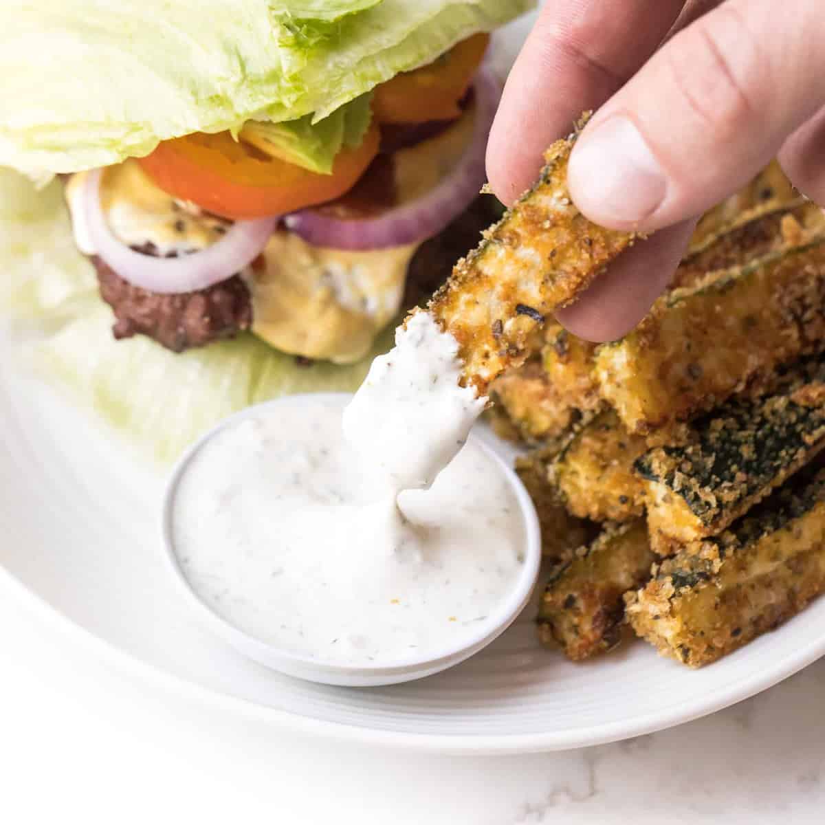 Hand dipping keto zucchini fries in ranch dressing on a white plate and white background with a lettuce wrapped burger on the plate