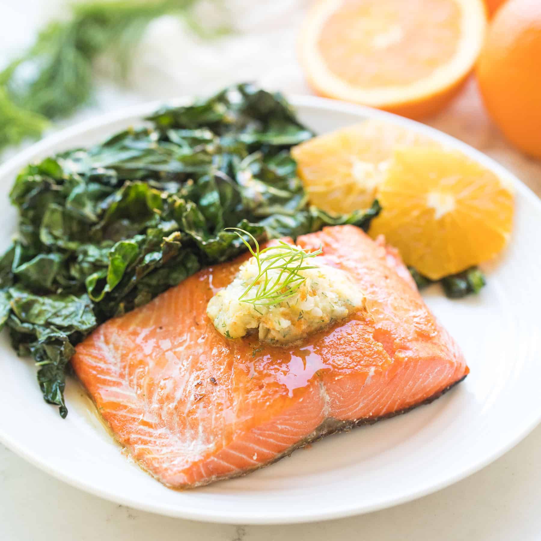 salmon on a white plate with butter on top next to peeled oranges and green kale salad