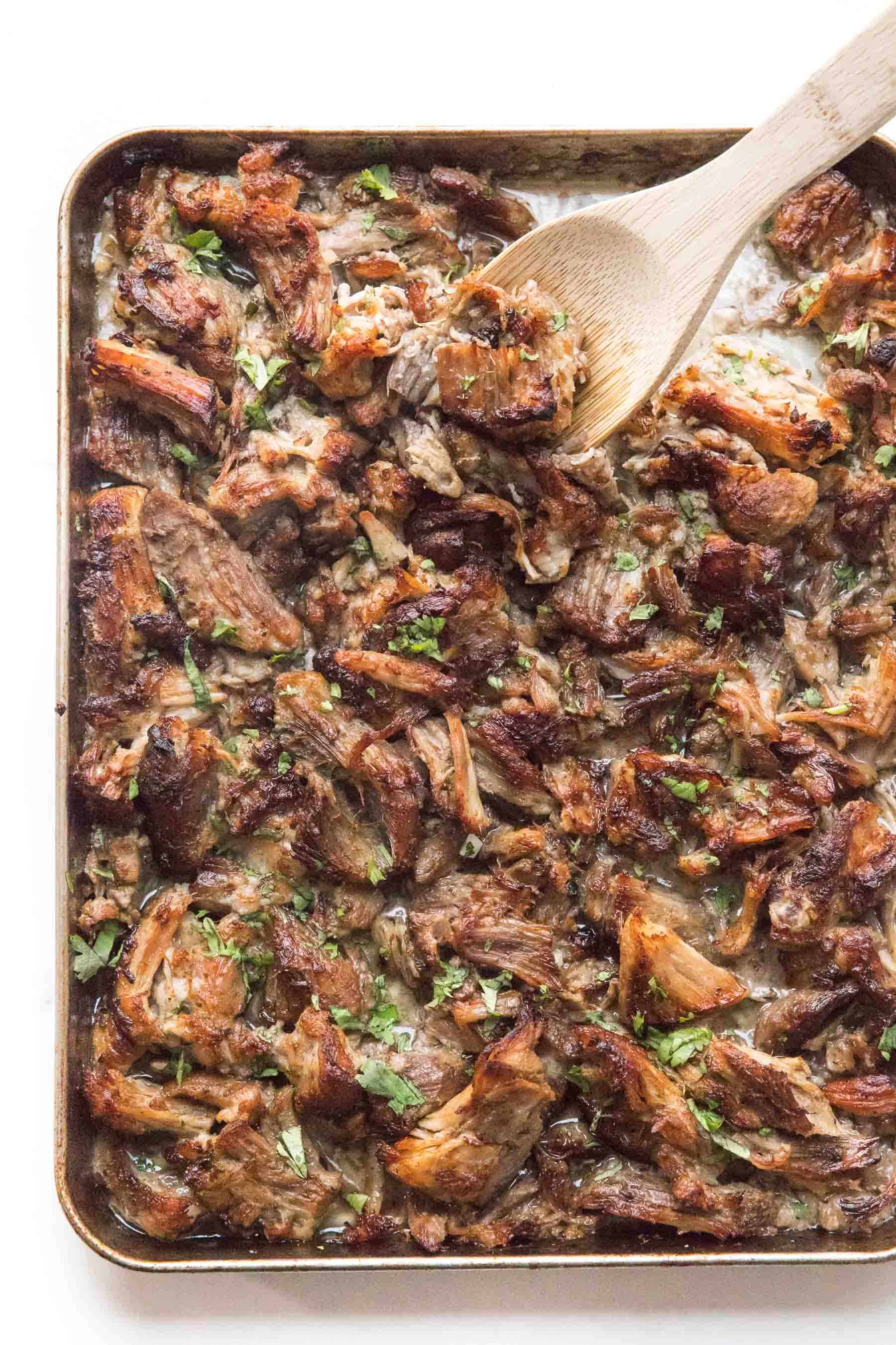 wooden spoon scooping crispy instant pot carnitas on a rimmed baking sheet sprinkled with cilantro