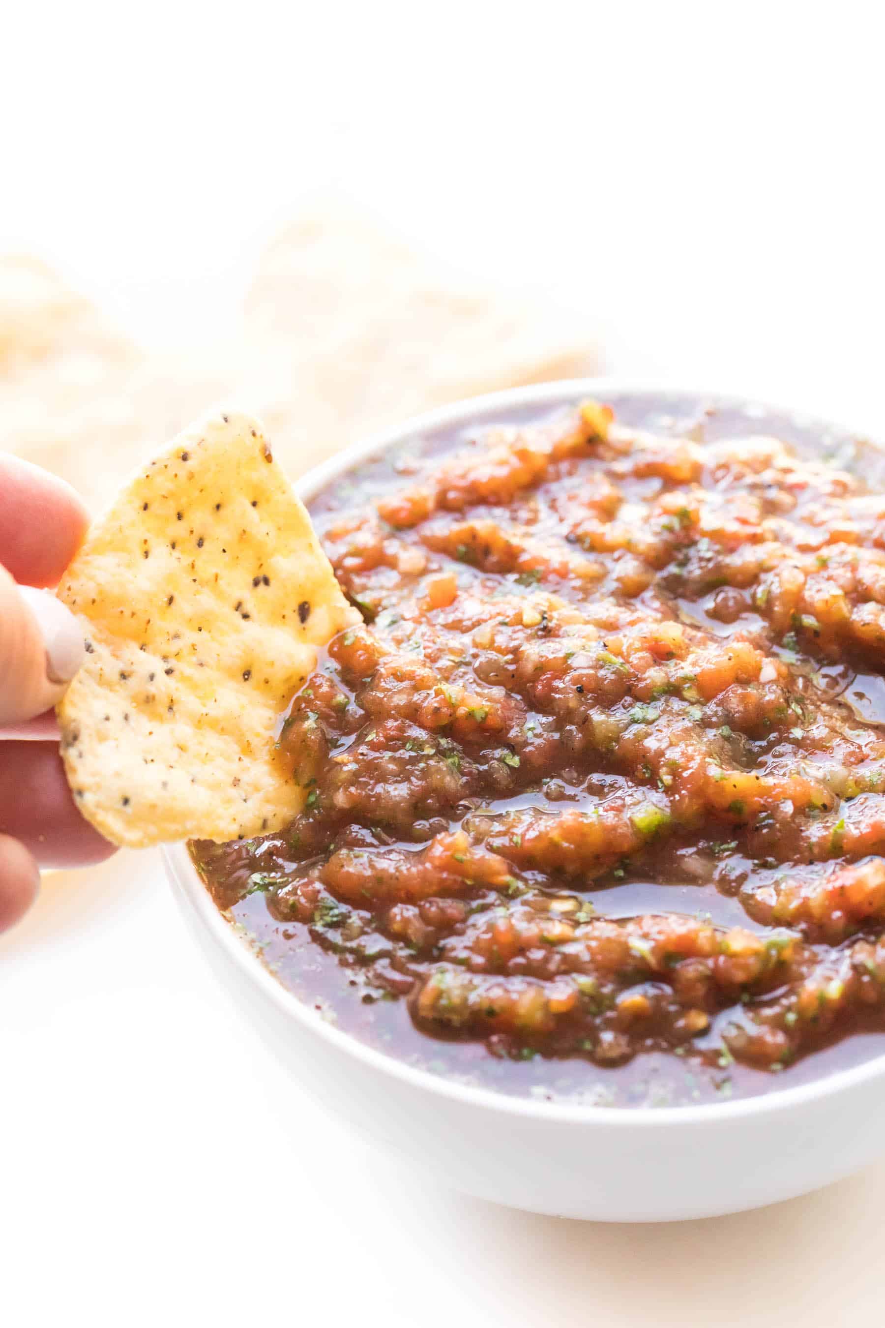 hand dipping a chip in homemade red salsa in a white bowl on a white background