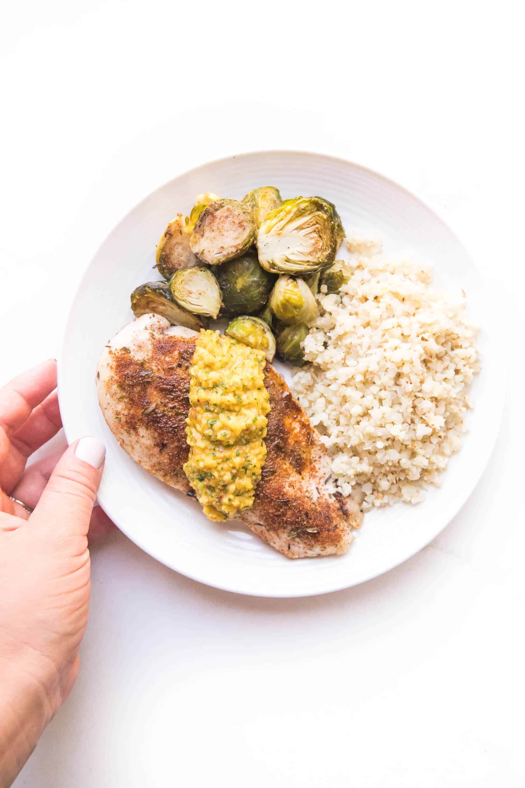 hand holding a plate with jamaican chicken topped with mango salsa with brussles sprouts and cauliflower rice on a white plate