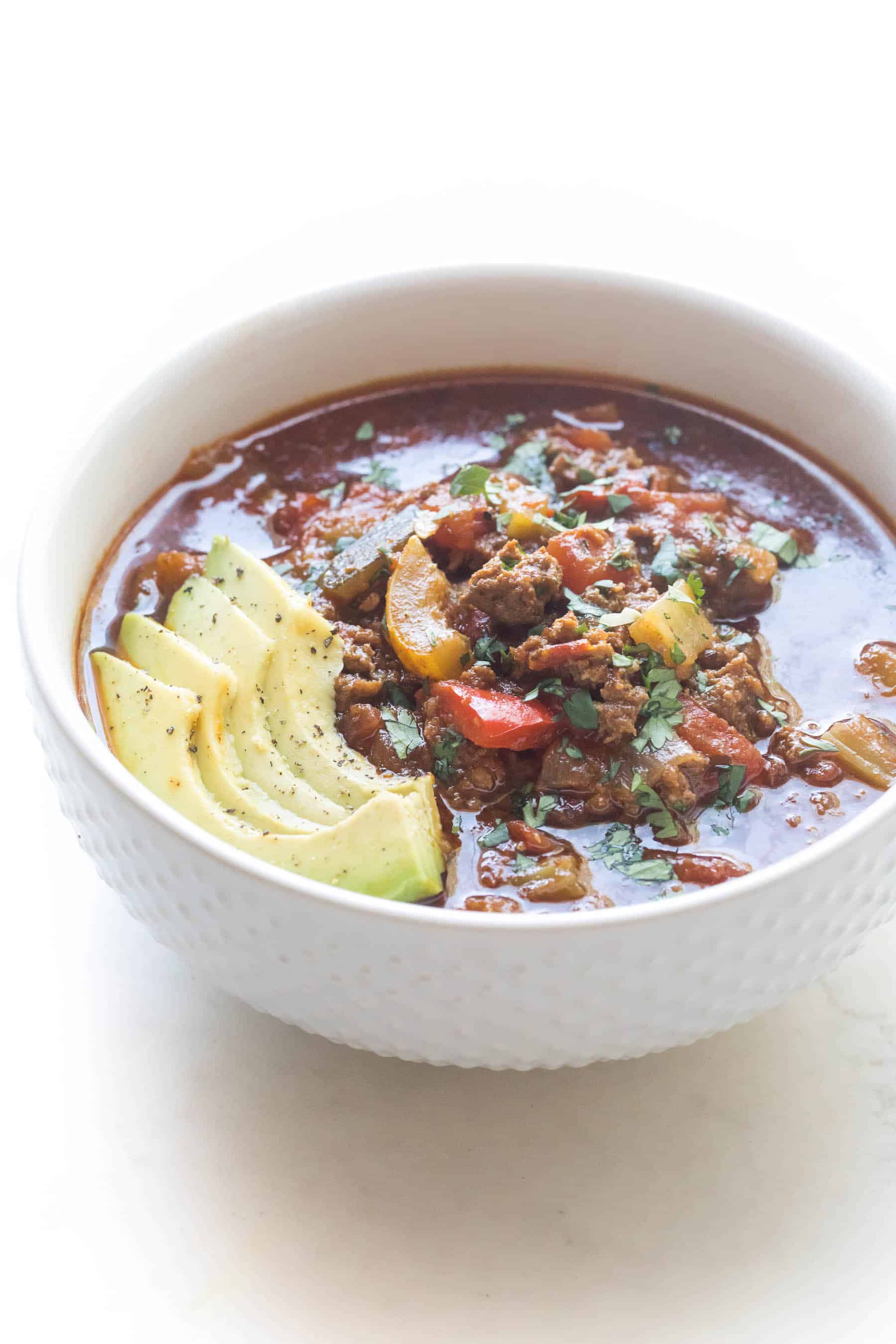 beef chili in a white bowl on a white background with avocado and cilantro