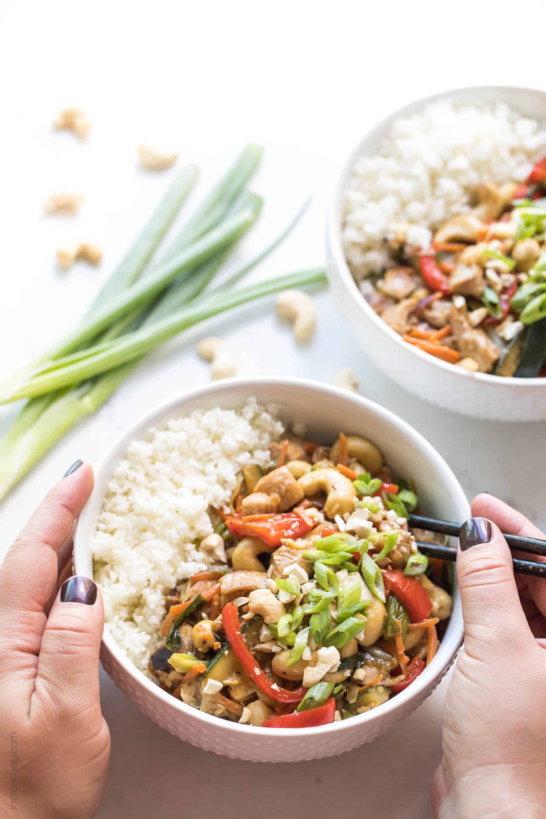 2 hands holding a bowl with chicken stir fry and cauliflower rice and chopsticks