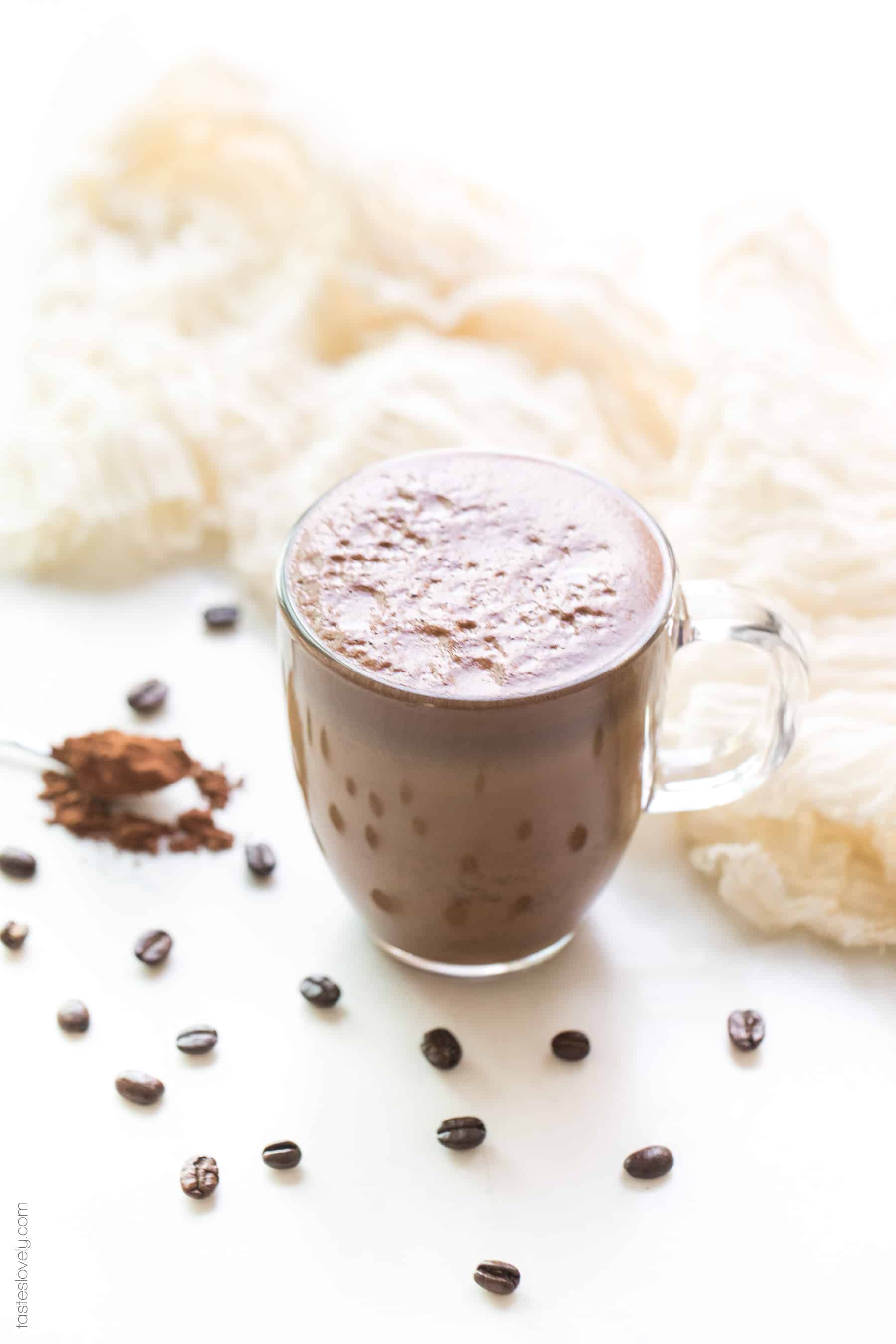 A mocha in a clear glass coffee cup on a white background with coffee beans and cocoa powder in the background