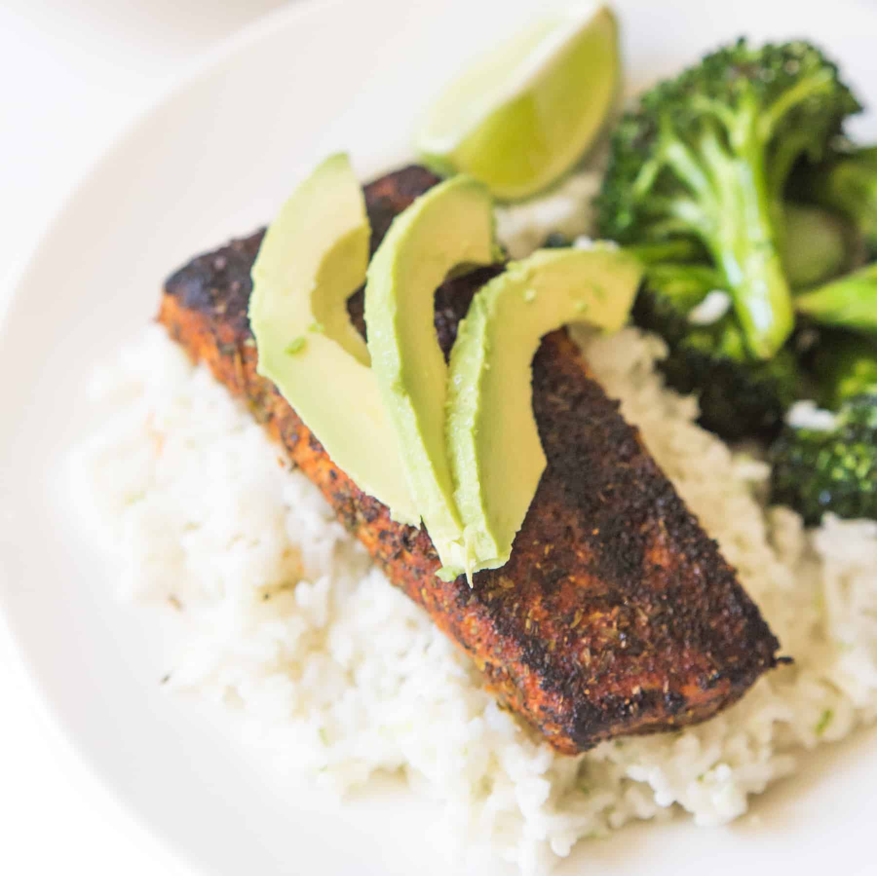 a piece of blackened Mahi Mahi over white rice on a plate topped with three avocado wedges and a lime wedge. 