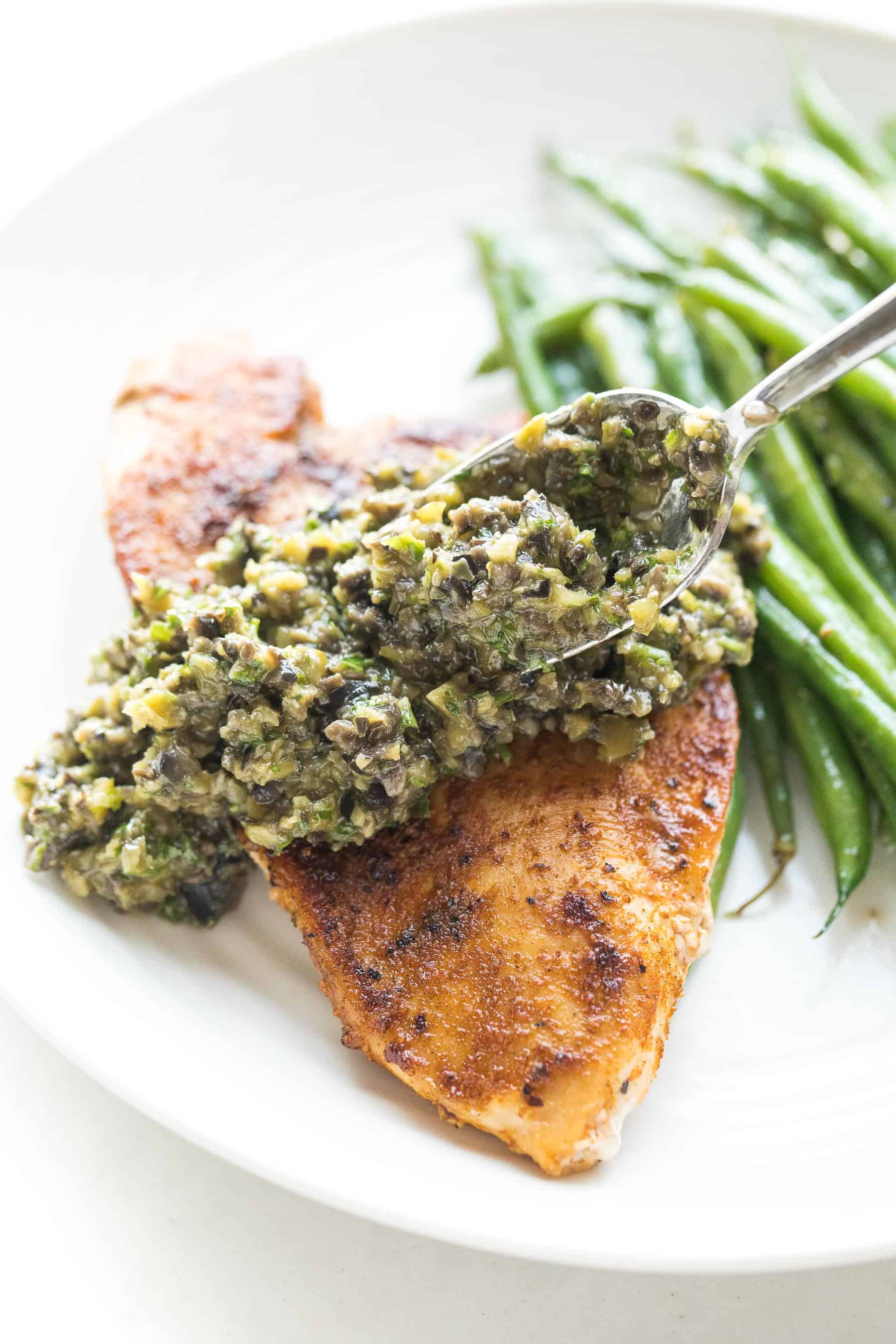 A spoon putting olive tapenade on top of chicken with green beans in the background