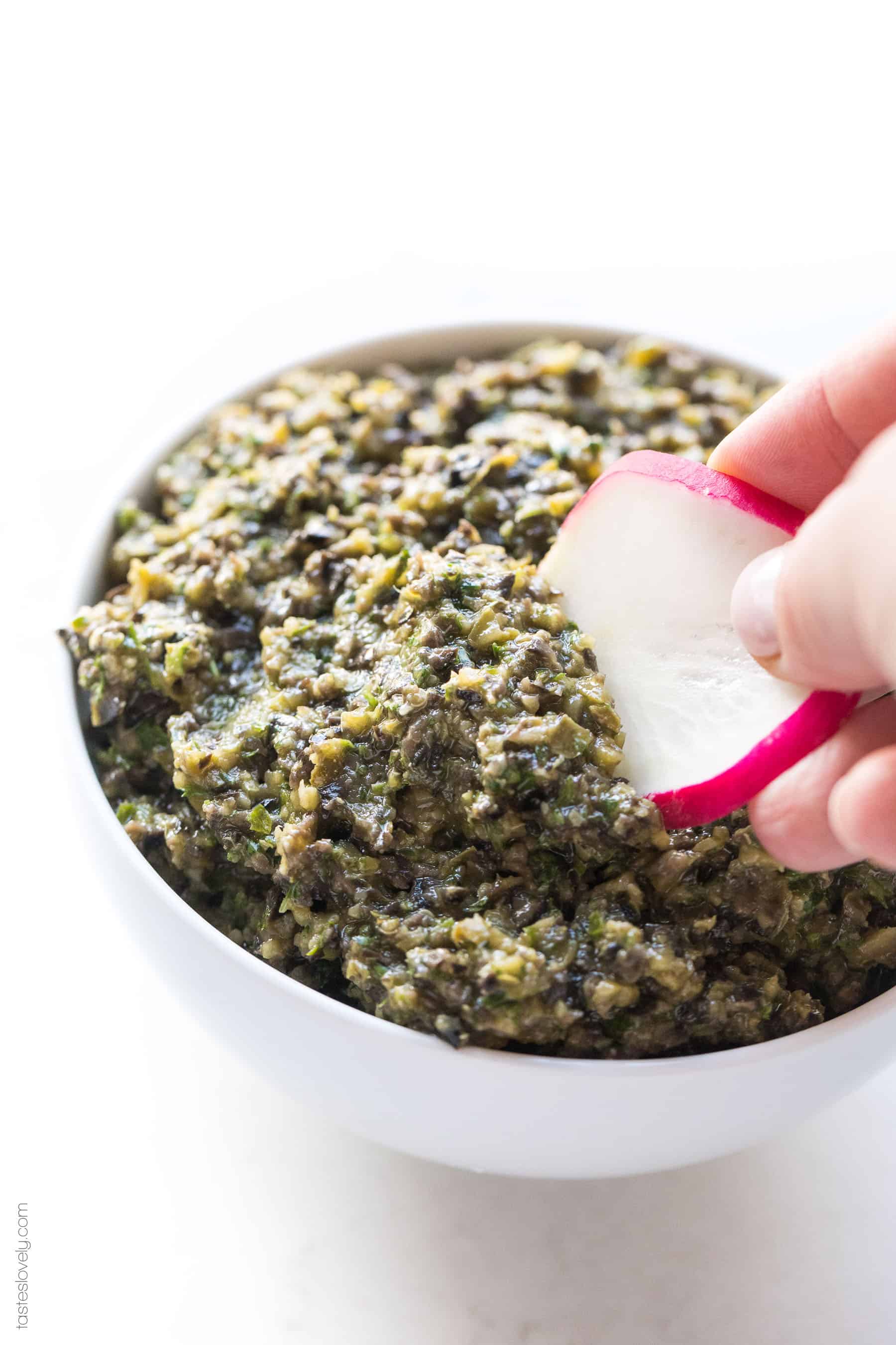 Radish being dipped into a white bowl of olive tapenade