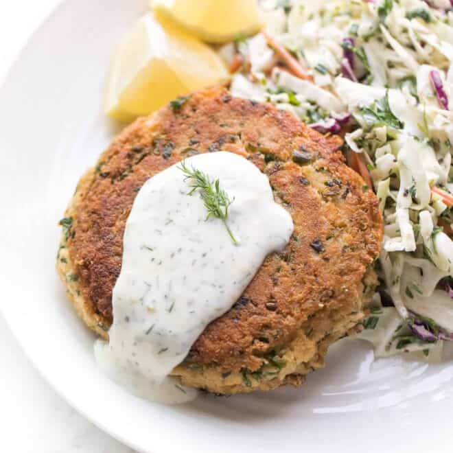 a salmon patty next to coleslaw and lemon wedges on a white plate