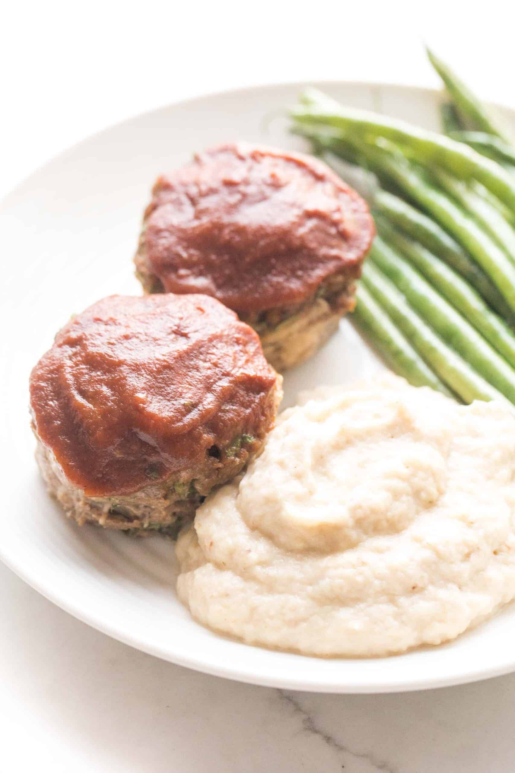 Mini meatloaf muffins topped with ketchup with green beans + mashed cauliflower + green beans on a white plate