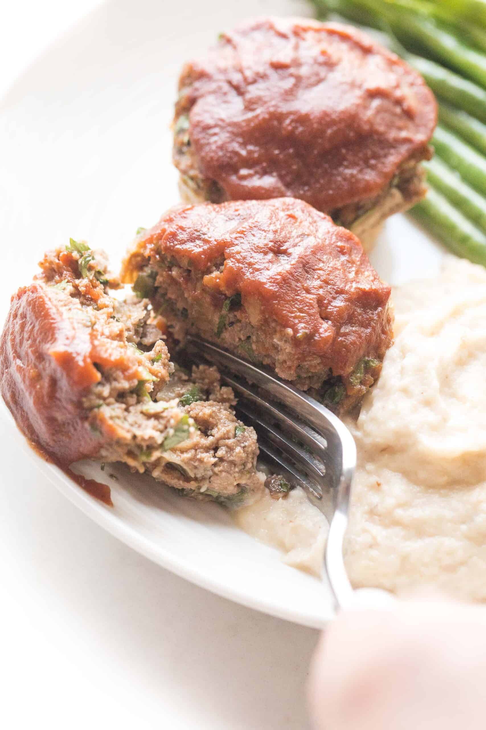knife cutting in mini meatloaf muffins topped with ketchup with green beans + mashed cauliflower + green beans on a white plate